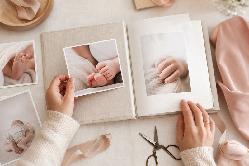 Parent arranging baby-detail photo prints in a linen-bound photo album with archival tissue paper and a soft ribbon