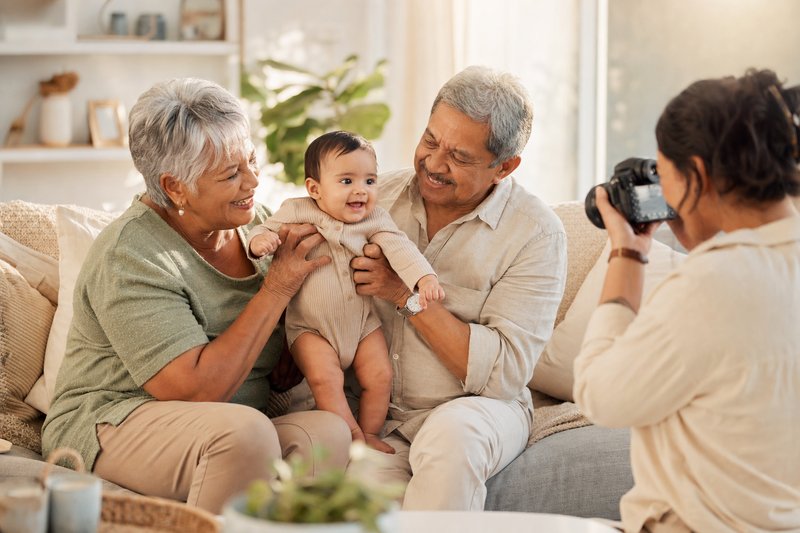 Grandparents joyfully holding a smiling infant while a family member photographs the candid living-room session