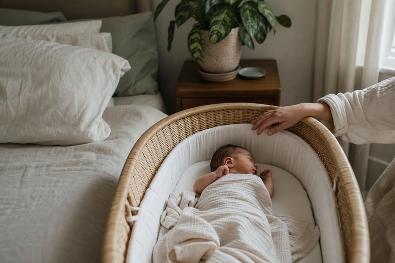 Newborn resting safely in a simple bassinet beside a bed in a calm sunlit home